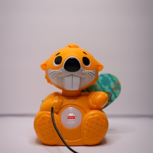 Pode incluir: Um brinquedo de castor laranja e verde com um botão luminoso e um cordão para puxar. O brinquedo é feito pela Fisher-Price.
