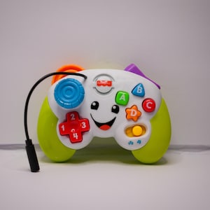 May include: A green and white toy video game controller with colorful buttons and a smiling face. The controller has a cord attached to it.