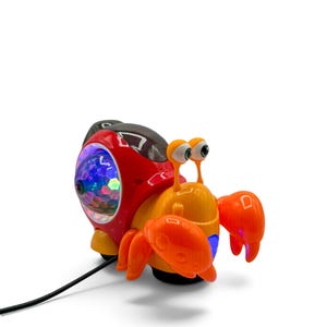 May include: A plastic crab toy featuring a bright red and orange body with two stalk eyes. The shell includes a circular, multi-colored light-up dome with a faceted surface. A black power cord extends from the base of the toy, which is positioned against a plain white background.