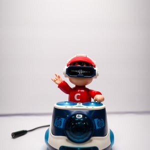 以下が含まれることがあります： 赤いシャツに白い「C」の文字が入ったおもちゃのDJフィギュア。赤と白のヘルメットとVRメガネを着用。フィギュアは、黒いケーブルが付いた青と白のDJブースの上にあります。