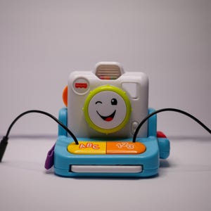 以下が含まれることがあります： レンズにウインクしているスマイリーフェイスが付いた青と白のオモチャのカメラ。カメラには「ABC」と「123」と書かれた2つのボタンがあります。カメラには黒いコードが2本付いています。