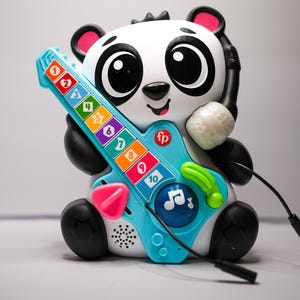 May include: A black and white panda toy holding a blue and green toy guitar with a pink button. The guitar has colorful keys with numbers 1-10 on them. The panda has a microphone attached to its ear.