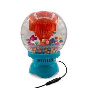 Könnte beinhalten: Ein Spielzeug mit einer transparenten Kunststoffblase mit einem orangefarbenen Oberteil und einem türkisfarbenen Sockel. Die Blase ist mit bunten Kugeln gefüllt. Ein schwarzes Kabel erstreckt sich von der Basis. Das Spielzeug ist zum Ausgeben der Kugeln konzipiert.