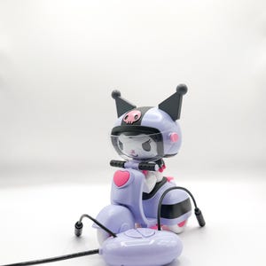 Peut inclure: Un scooter jouet violet clair avec une télécommande assortie. Le scooter présente un personnage portant un casque avec des oreilles noires et un motif de crâne. Le scooter a un cœur rose à l'avant et des rayures noires et blanches sur le côté.
