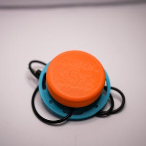 May include: An orange and blue device with a black cord. The orange top has embossed text. The device is circular with a blue ring around the orange top. The black cord has a loop at the end.