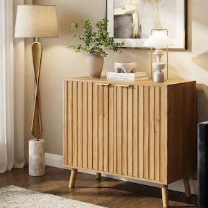 May include: A light oak wood cabinet with vertical slats on the two doors, featuring thin gold metal handles and four tapered wooden legs. The cabinet top holds a ceramic vase with greenery, a small stone bowl, and two stacked white books.