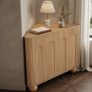 May include: A light oak corner cabinet featuring two doors with arched recessed panels and rounded ball feet. On top sits a white marble-based lamp with a pleated shade, a closed book with a brown spine, and a small vase containing dried foliage.