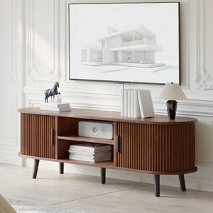 May include: A dark wood media console with a rounded, fluted design, featuring two side cabinets with vertical slats, a central open shelf containing a white rectangular device, and a stack of books, supported by four tapered black legs.