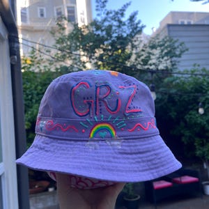 May include: A purple bucket hat with the word "GRZ" in colorful lettering. The hat features a rainbow and other decorative elements. The hat is held up by a hand.