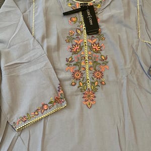 以下が含まれることがあります： ピンク、オレンジ、緑、黄色の花の刺繍が施された水色のチュニック。チュニックは長袖で、ネックライン、袖口、中央に金色のトリムが付いています。「cast & crew」というテキストの黒いタグが取り付けられています。