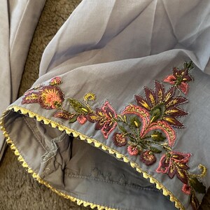 以下が含まれることがあります： ピンク、緑、金色の糸で施された、繊細な花の刺繍が施された、明るいグレーの衣服の袖のクローズアップ。縁は金色のスカラップボーダーでトリミングされています。生地は柔らかく、流れるように見えます。