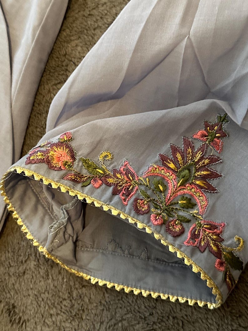 以下が含まれることがあります： ピンク、緑、金色の糸で施された、繊細な花の刺繍が施された、明るいグレーの衣服の袖のクローズアップ。縁は金色のスカラップボーダーでトリミングされています。生地は柔らかく、流れるように見えます。