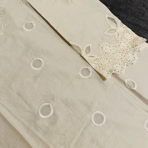 May include: A white fabric with a delicate lace design. The lace features a floral pattern with small circles throughout.