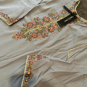 以下が含まれることがあります： 薄いグレーの刺繍入りチュニックとパンツのセット。チュニックはピンク、オレンジ、緑の色合いの花柄刺繍が施され、ネックラインと袖にはゴールドのトリムが施されています。「cas&crew」と書かれたタグが付いています。