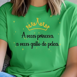 Puede incluir: Una camiseta de manga corta de color verde brillante con un gráfico de una corona amarilla en la parte superior y el texto negro "A veces princesa, a veces gallo de pelea." El diseño incluye pequeñas estrellas amarillas de cuatro puntas.