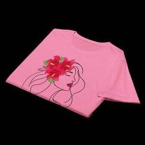 Puede incluir: Camiseta rosa con un dise&ntilde;o de perfil de mujer, pelo largo y un tocado floral. Las flores son rojas con hojas verdes, y la mujer tiene labios rojos. El dise&ntilde;o est&aacute; en la parte delantera de la camiseta.