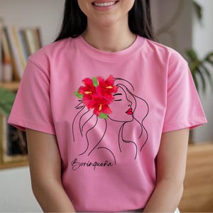 Puede incluir: Camiseta rosa con un dibujo lineal de una mujer adornada con flores de hibisco rojas y pintalabios rojo. La palabra "Boricua" est&aacute; escrita en cursiva en la parte inferior. La camiseta es de un material suave.
