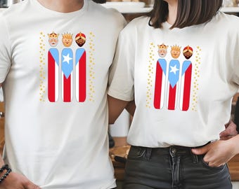May include: White t-shirts featuring a design with the Puerto Rican flag and three cartoon figures wearing crowns. The design includes red, white, and blue colors, with gold stars. The shirts are worn by two people.