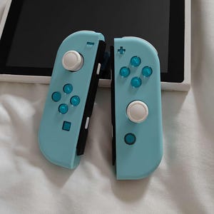 May include: A handheld video game console featuring a white screen unit and two detachable light blue controllers. The controllers have white analog sticks and translucent blue directional buttons, including a plus sign and four circular buttons.