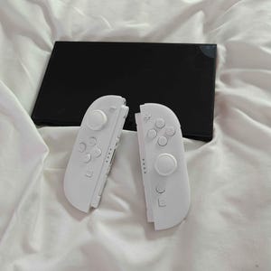 May include: A black games console with two detached white controllers. The controllers have buttons and joysticks. The console and controllers are on a white, creased surface.