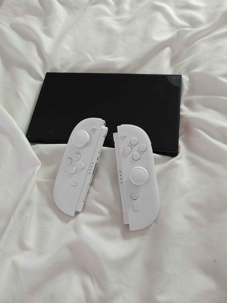 May include: A black games console with two detached white controllers. The controllers have buttons and joysticks. The console and controllers are on a white, creased surface.