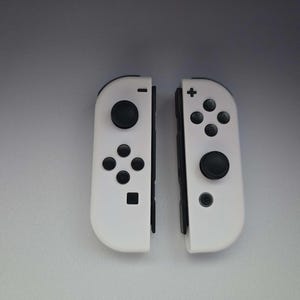 May include: Two white game controllers with black buttons and directional pads. The controllers are designed for a handheld gaming console. Each controller has a joystick and several buttons for gameplay.