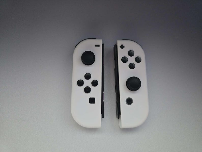 May include: Two white game controllers with black buttons and directional pads. The controllers are designed for a handheld gaming console. Each controller has a joystick and several buttons for gameplay.