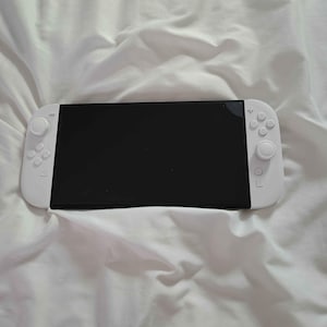 May include: A white handheld games console with a black screen, and white control pads on either side. The console is on a white, creased fabric surface. Buttons and joysticks are visible on the console.