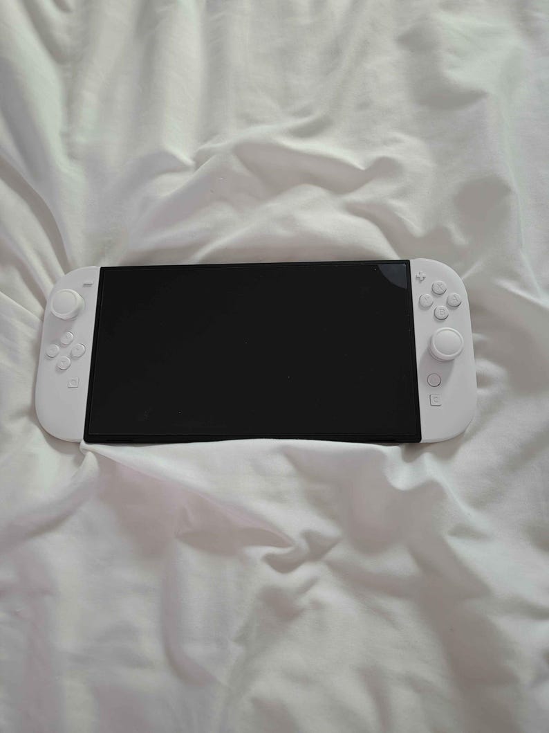 May include: A white handheld games console with a black screen, and white control pads on either side. The console is on a white, creased fabric surface. Buttons and joysticks are visible on the console.