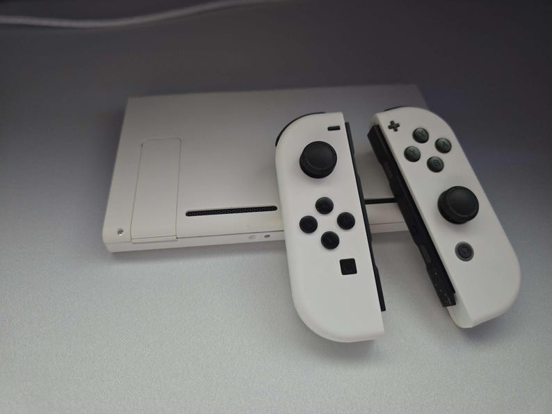 May include: A white gaming console with two detached white controllers. The controllers have black buttons and joysticks. The console is rectangular with a slot on the side.
