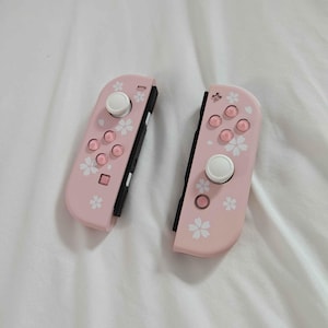 May include: Two pink game controllers with white buttons and a floral design. The controllers have a black side and are designed for a gaming console. The controllers are on a white surface.