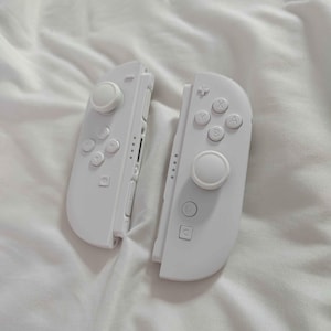 May include: Two white Nintendo Switch Joy-Con controllers. Each controller has a joystick, buttons, and a directional pad. The controllers are resting on a white, textured surface.