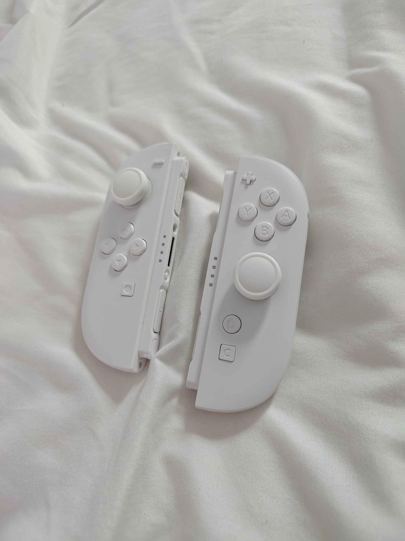 May include: Two white Nintendo Switch Joy-Con controllers. Each controller has a joystick, buttons, and a directional pad. The controllers are resting on a white, textured surface.