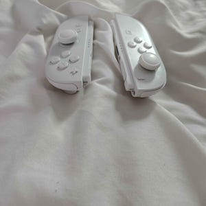 May include: Two white game controllers, each with a joystick, buttons, and directional pad. The controllers are on white, creased fabric. The controllers are designed for a gaming console.