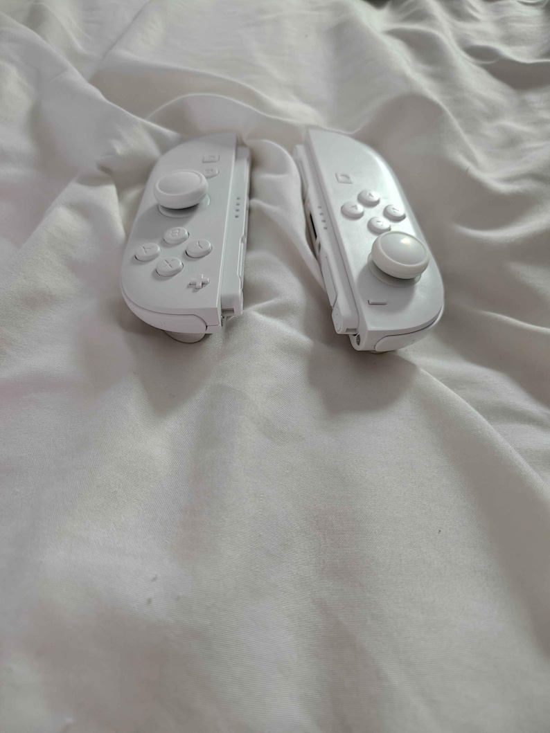 May include: Two white game controllers, each with a joystick, buttons, and directional pad. The controllers are on white, creased fabric. The controllers are designed for a gaming console.