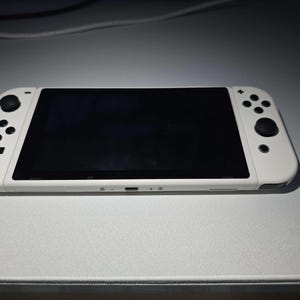 May include: A white handheld gaming console with a black screen and black control buttons. The console is lying on a light gray surface. The console has a sleek, modern design.