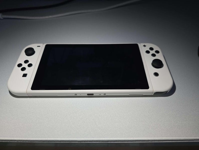 May include: A white handheld gaming console with a black screen and black control buttons. The console is lying on a light gray surface. The console has a sleek, modern design.