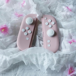 Könnte beinhalten: Ein Paar matte, pastellrosa Videospiel-Controller mit weißen Analogsticks, weißen Richtungstasten und weißen Aktionstasten mit der Beschriftung X, Y, A und B, die auf einem weißen, gerüschten Stoff mit verstreuten rosa und weißen Blüten liegen.