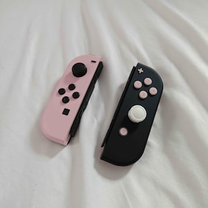 May include: Two Nintendo Switch Joy-Con controllers. One is light pink with black buttons and a black joystick. The other is black with pink buttons and a white joystick. Both controllers are lying on a white surface.