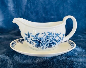 May include: A white porcelain gravy boat and saucer set. The boat and saucer feature a blue floral design. The gravy boat has a handle and a pouring spout. The set is displayed on a blue backdrop, typical of a dining setting.