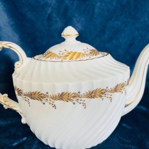 May include: White porcelain teapot with a gold leaf design and a spiral pattern. The teapot has a curved handle and spout, and a lid with a finial. The gold leaf design is on the lid and around the body of the teapot.