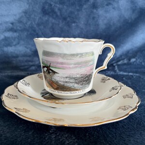 May include: A white teacup and saucer set with gold edging. The teacup has a scenic image of a sunset over the sea, with text reading "Sunset, Qualicum Beach, V.I.B.C." The set includes a small and a larger plate.