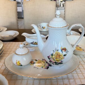 May include: A white porcelain tea set with a floral rose design. The set includes a teapot, sugar bowl, serving tray, and various plates and cups. Gold trim accents the edges of the pieces. The tea set is arranged on a table with a patterned tablecloth.