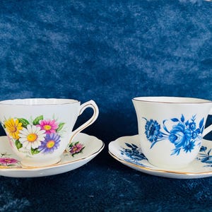 May include: Two white teacups and saucers. One cup features a floral design with yellow, pink, and purple flowers. The other cup has blue floral patterns. Both have gold trim.