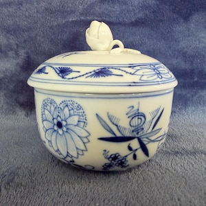 May include: A white porcelain sugar bowl with a rounded body and a matching lid featuring a sculpted rosebud finial. The exterior is decorated with intricate blue floral patterns, including stylized blossoms, leaves, and vines painted in a delicate style.