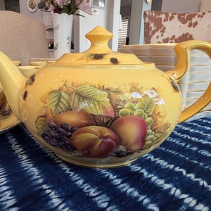 May include: A yellow ceramic teapot with a gold handle and lid. The teapot is decorated with a painted design of fruit, including grapes, peaches, and apples. The teapot is part of a tea set.