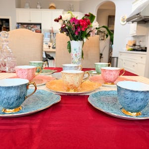 May include: A set of pastel-coloured teacups and saucers on a red tablecloth. The teacups, in shades of pink, blue, green, and yellow, have gold detailing. The oval saucers match the cups. A vase of flowers is visible in the background, adding to the scene.