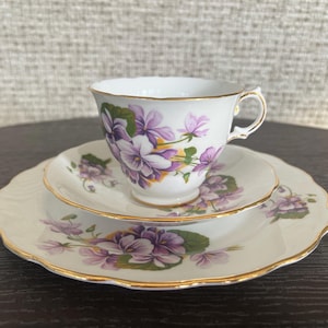May include: A white teacup, saucer, and side plate set. The set features a purple violet floral design with green leaves and gold trim. The edges of the plates are scalloped.