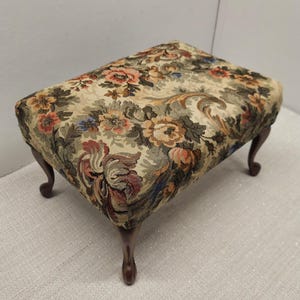 May include: A rectangular footstool featuring a woven tapestry fabric upholstery with a multi-colored floral pattern in shades of orange, green, blue, and cream. The footstool is supported by four dark brown wooden legs with a curved cabriole design.