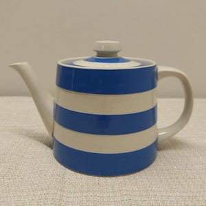 May include: A ceramic teapot featuring a cylindrical body with horizontal blue and white stripes. The teapot includes a matching lid with a small knob handle, a curved spout on the left side, and a curved handle on the right side, all in white.